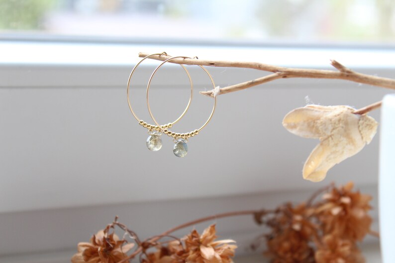 May include: Gold hoop earrings with small, clear green gemstone drops hanging from them. The earrings are displayed on a branch.