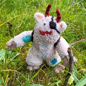Op de afbeelding: Een wit, pluche, handgemaakt speelgoedmonster met rode hoorns, een zwart ooglapje en blauwe accenten. Het speelgoed zit in een bed van groen gras.