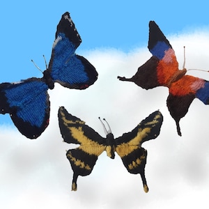 May include: Three knitted butterflies against a blue sky with white clouds. The butterflies are blue and black, orange and black, and yellow and black.