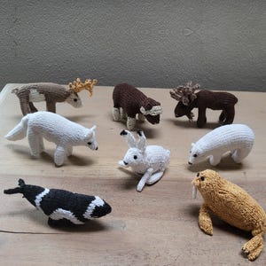 Pode incluir: Uma coleção de figuras de animais de malha, incluindo uma rena, um urso polar, um lobo, um boi almiscarado, um alce, um coelho, uma baleia e uma morsa. As figuras são todas brancas, marrons ou pretas, e estão dispostas numa superfície de madeira.