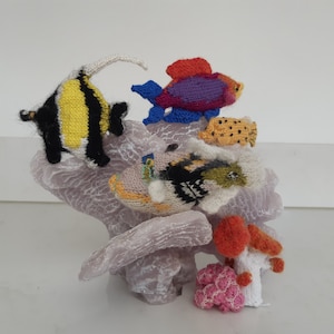 May include: Knitted fish figurines on a coral reef base. The fish are yellow and black, purple and blue, and brown and white. There are also two coral pieces, one pink and one orange.