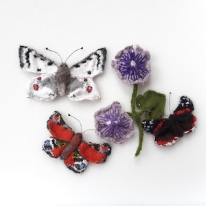以下が含まれることがあります： 4匹のニットの蝶と花。2匹の蝶は白地に黒と赤のアクセント。1匹の蝶は赤地に黒と黄色のアクセント。もう1匹の蝶は黒地に白のアクセント。花は紫と白です。