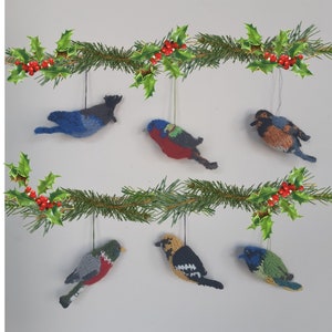 May include: A collection of six knitted bird ornaments hanging from a garland of green holly branches with red berries. The birds are all different colors and patterns, including blue, red, green, yellow, and brown.