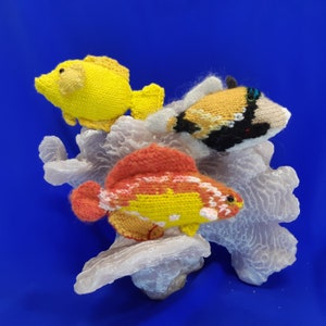 May include: Three knitted fish figurines on a white coral reef. The fish are yellow, orange, and black with white accents. The fish are sitting on a white coral reef.