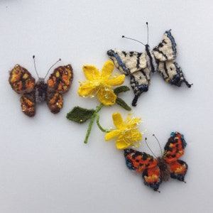 May include: Hand-knitted butterfly and flower embellishments. The butterflies feature orange, brown, and cream-colored wings with beaded accents. A yellow flower with green leaves and stems is also present. These decorative items are perfect for craft projects.