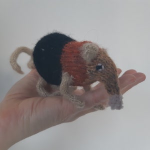 May include: A hand-knitted plush toy of a brown and black opossum with a long tail. The opossum is wearing a red and orange knitted sweater.