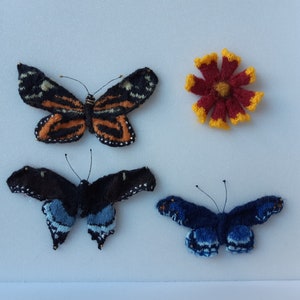 May include: Three knitted butterflies and a knitted flower. The butterflies are black and blue, black and white, and orange and black. The flower is red with yellow edges.