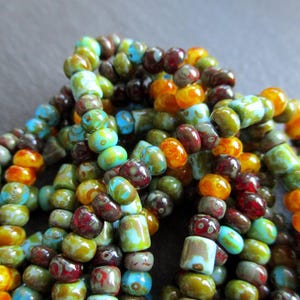Pode incluir: Um close-up de um monte de contas de vidro coloridas. As contas são em tons de laranja, amarelo, verde, azul e marrom. Todas são pequenas e redondas, e estão dispostas em um padrão aleatório.