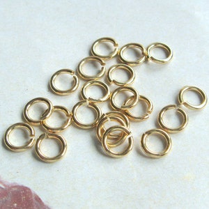 Op de afbeelding: Een verzameling gouden springringen, een soort sieraadbevestiging, die wordt gebruikt om sieraadcomponenten te verbinden. De ringen zijn van verschillende groottes en zijn in een willekeurig patroon gerangschikt.