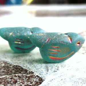 Può includere: Due perline a forma di uccello color turchese con dettagli iridescenti. Le perline hanno una superficie strutturata e linee incise che suggeriscono ali e becco. Per la creazione di gioielli.
