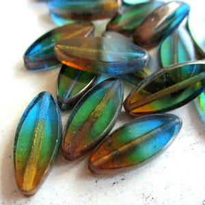 Könnte beinhalten: Eine Sammlung ovaler Glasperlen mit einem lebendigen Farbverlauf. Die Perlen wechseln von Blau- und Grüntönen zu Bernstein und Braun. Jede Perle hat einen zentralen, metallisch aussehenden Streifen.
