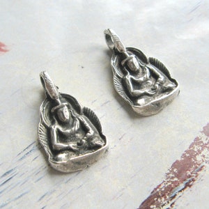 Puede incluir: Dos colgantes de plata con una representación detallada de una figura de Buda sentado. Los colgantes tienen una superficie ligeramente texturizada y un bucle en la parte superior para sujetarlos a una cadena.