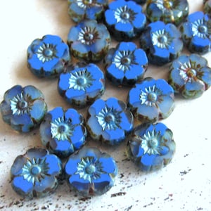 Könnte beinhalten: Eine Sammlung von blauen Glasperlen in Blumenform mit einem silbernen, irisierenden Finish. Die Perlen sind in einem Halbkreis auf einer weißen Oberfläche angeordnet.