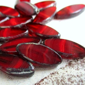 Może przedstawiać: Zbliżenie na kilka owalnych, głęboko czerwonych szklanych koralików. Koraliki mają błyszczące wykończenie z ciemną, prawie czarną obwódką. Koraliki są rozrzucone na jasnej, teksturowanej powierzchni, tworząc atrakcyjny wizualnie układ.