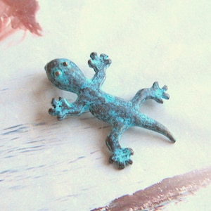 May include: A turquoise blue metal gecko figurine with a textured finish. The gecko is posed with its legs outstretched and its tail curled.