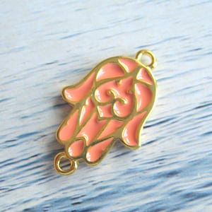 Może przedstawiać: Złoty charm z wzorem ręki Hamsy w różowym szkliwie. Charm ma dwa kółka do zawieszania na biżuterii.