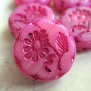 Könnte beinhalten: Nahaufnahme mehrerer rosa Knöpfe mit floralem Design. Jeder Knopf hat ein erhabenes Blumen- und Blättermuster. Die Knöpfe sind in einem leuchtenden Rosaton gehalten und wirken leicht durchscheinend. Die Knöpfe sind rund und leicht gewölbt.