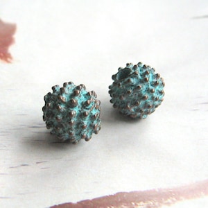 Può includere: Due perline di metallo blu turchese con una superficie testurizzata e spinosa. Le perline sono rotonde e hanno un aspetto rustico e invecchiato.