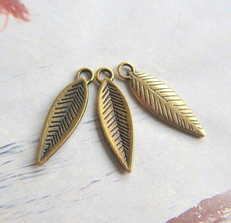 K&ouml;nnte beinhalten: Drei goldfarbene Metall-Bl&auml;tter-Anh&auml;nger mit strukturiertem Design. Die Bl&auml;tter sind l&auml;nglich und haben eine spitze Spitze.