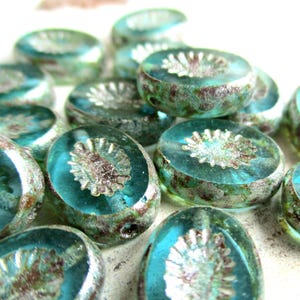 Pode incluir: Uma coleção de contas de vidro azul-turquesa com um desenho floral no centro. As contas têm uma forma ligeiramente oval e um aspeto texturizado e envelhecido. Adequadas para a criação de joias e outros projetos de artesanato.