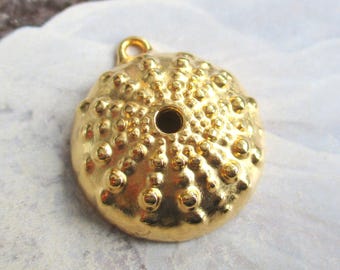 May include: A gold-colored, round pendant with a textured surface, resembling a sea urchin. The pendant has a small loop at the top for hanging and a central hole. The surface is covered in small, raised bumps. Jewelry.