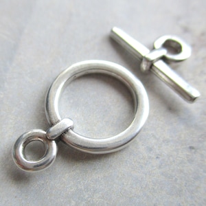 Könnte beinhalten: Silberner Toggle-Verschluss mit einem runden Ring und einer Stange mit einer Schlaufe. Der Verschluss ist ein beliebtes Schmuckelement für Halsketten, Armbänder und andere Schmuckprojekte.