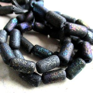 Könnte beinhalten: Nahaufnahme eines Strangs dunkler, zylindrischer Perlen. Die Perlen haben eine strukturierte, gesprenkelte Oberfläche mit schillernden Farbvariationen in Blau, Lila und Grün. Die Perlen sind aufgefädelt und bereit für die Schmuckherstellung.