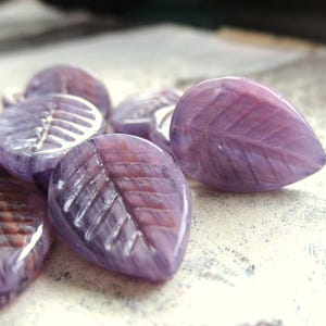 Peut inclure: Une collection de petites perles en verre en forme de feuille, dans des tons marbrés de lavande et de violet. Chaque perle présente un motif de nervures en relief gravé sur la surface, créant un aspect texturé avec de fines stries brun-rouge.