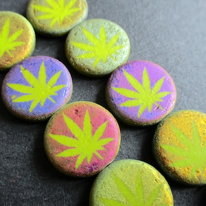 Può includere: Sette pietre rotonde, iridescenti, dipinte con un motivo a foglia di marijuana verde. Le pietre sono in tonalità di viola, blu, verde e oro.