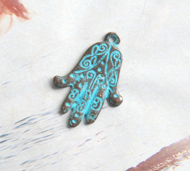 May include: A turquoise blue metal charm in the shape of a hand with intricate designs. The charm has a loop at the top for attaching to jewelry or other crafts.