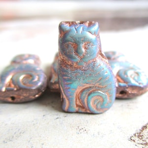 May include: A group of iridescent blue and copper-toned cat-shaped beads. The beads feature a textured finish with etched details, including swirls and facial features. These cat beads are a unique craft supply.