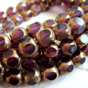 Pode incluir: Close-up de um fio de contas. As contas são uma mistura de ouro e roxo, com um brilho metálico. Cada conta tem um design facetado com um centro roxo escuro. As contas são redondas e parecem ser feitas de vidro ou material semelhante.