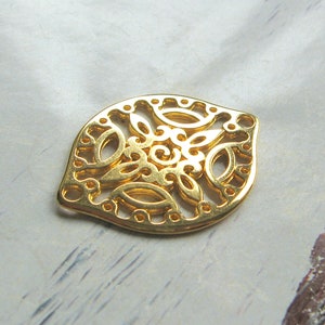 Może przedstawiać: Złoty, ażurowy metalowy wisiorek z misternym kwiatowym wzorem. Wisiorek ma kształt liścia z zaokrąglonym dołem i spiczastym wierzchołkiem. Projekt zawiera wirujące wzory i owalne wycięcia.