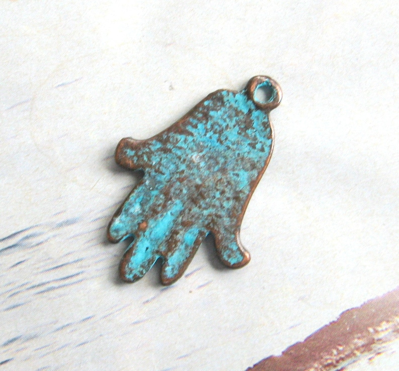 May include: A turquoise blue and brown metal charm in the shape of a hamsa hand. The charm has a distressed finish and a small loop at the top for attaching to a necklace or bracelet.