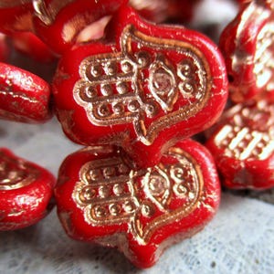 Peut inclure: Perles de verre rouges avec des charmes de main de hamsa dorés. Les perles sont enfilées pour former un collier ou un bracelet.