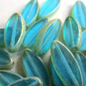Peut inclure: Gros plan de plusieurs perles de forme ovale. Les perles sont d'un bleu-vert translucide avec un bord doré. Les perles ont une texture côtelée et sont dispersées sur une surface claire. Ces perles conviennent à la fabrication de bijoux.
