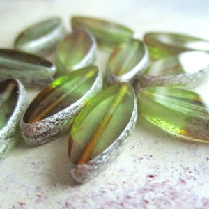 May include: A group of green glass beads with silver accents. The beads are oval shaped and have a translucent appearance. The beads are arranged in a random pattern.