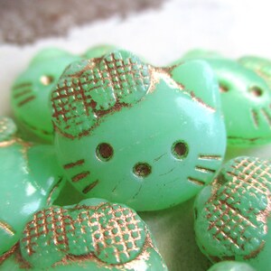 Może przedstawiać: Zbiór miętowo-zielonych koralików w kształcie kota z postarzanym, metalicznym złotym wykończeniem. Koraliki mają wytłoczone detale, w tym kokardkę i rysy twarzy. Te ozdobne koraliki są idealne do rękodzieła i tworzenia biżuterii.