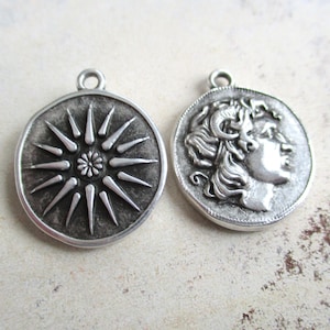Könnte beinhalten: Zwei silberfarbene Metallanhänger. Ein Anhänger zeigt ein Sonnenstrahl-Design mit einem zentralen Blumenelement. Der andere Anhänger zeigt das Profil einer Person mit lockigem Haar, das einer antiken Münze ähnelt.