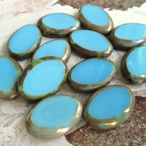 Puede incluir: Una colección de cuentas de vidrio ovaladas con un esmalte azul claro y un borde plateado metálico. Las cuentas tienen un aspecto desgastado y vintage.