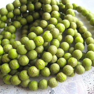 Pode incluir: Um fio de contas de vidro verde com uma superfície texturizada. As contas são redondas e têm um brilho sutil.