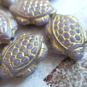 Op de afbeelding: Een close-up van verschillende paarse glazen schildpad kralen met gouden accenten. De kralen zijn gedetailleerd met een textuur van een schildpad schild.