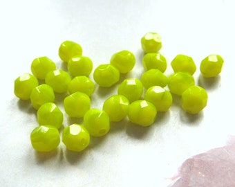 May include: A collection of faceted, lime green beads. The beads have a rounded shape with multiple flat surfaces, creating a sparkling effect. The beads are scattered across a white surface.