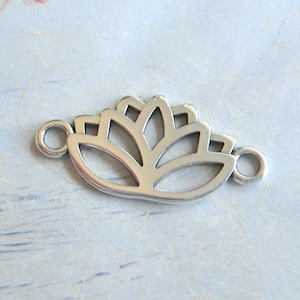 Może przedstawiać: Srebrny metalowy urok w kształcie kwiatu lotosu z dwoma kółkami do zawieszenia. Urok jest delikatny i ażurowy.