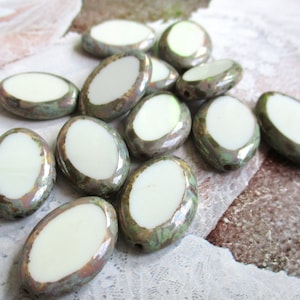 Puede incluir: Un grupo de cuentas de cerámica ovaladas con un centro blanco y un esmalte verde iridiscente. Las cuentas tienen una forma ligeramente irregular y una superficie texturizada.