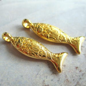 Può includere: Due ciondoli a forma di pesce dorati con un design intricato e testurizzato. I ciondoli hanno un anello nella parte superiore per essere attaccati a gioielli o altri oggetti artigianali.
