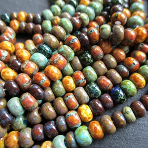 Pode incluir: Close-up de uma coleção de contas coloridas em forma de barril. As contas são uma mistura de cores, incluindo laranja, castanho, verde e azul. Algumas contas têm padrões salpicados, enquanto outras têm uma cor sólida. As contas são enfiadas.