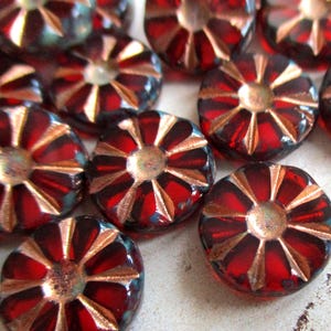 Könnte beinhalten: Nahaufnahme mehrerer roter Glasperlen mit einem blütenartigen Design. Jede Perle hat einen zentralen metallisch goldfarbenen Kreis mit strahlenförmigen goldfarbenen Linien. Die Perlen sind rund und leicht gewölbt.