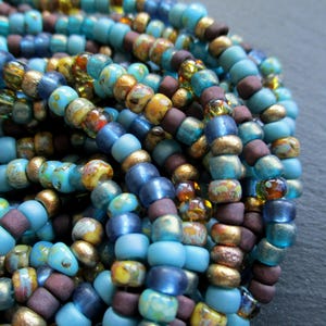 Könnte beinhalten: Nahaufnahme einer Sammlung farbenfroher Perlen in Türkis-, Blau-, Braun- und Goldtönen. Die Perlen haben verschiedene Formen und Größen und erzeugen ein strukturiertes und geschichtetes Aussehen. Die Perlen sind wahrscheinlich für die Schmuckherstellung oder das Basteln gedacht.