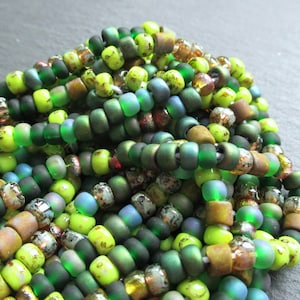 Könnte beinhalten: Nahaufnahme eines Haufens grüner, gelber und brauner Glasperlen. Die Perlen sind in einem zufälligen Muster angeordnet und haben alle unterschiedliche Größen. Die Perlen sind perfekt für die Schmuckherstellung oder andere Bastelprojekte.